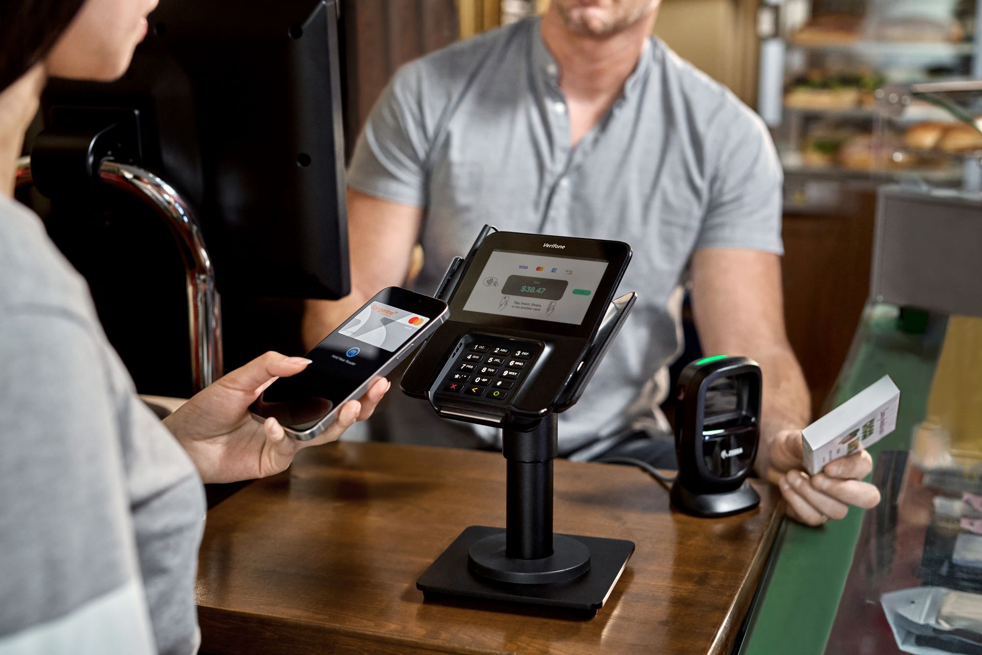 Customer using smartphone for contactless payment at a cashier counter with a Verifone card reader displaying $38.47.